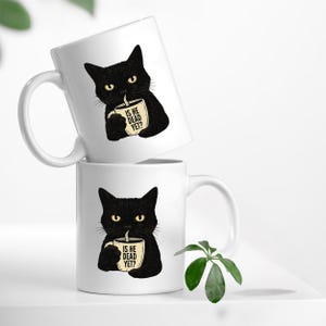 May include: Two white ceramic mugs stacked on top of each other. Each mug features a black cat illustration holding a coffee cup. The coffee cup has the text "IS HE DEAD YET?" printed on it.