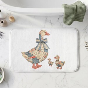 May include: A rectangular white bath mat featuring a central graphic of a large goose and two small ducklings. The birds are decorated with a patchwork pattern of blue, red, and white floral and gingham fabrics, with a blue gingham bow on the goose.