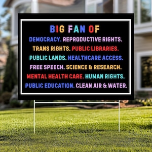 May include: A black yard sign with a white border and text in rainbow colours. The text reads "BIG FAN OF" followed by a list of values including democracy, reproductive rights, and public education. The sign is supported by two white metal stakes.