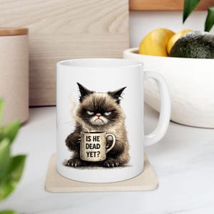 May include: White ceramic mug featuring a grumpy cat illustration holding a coffee cup. The mug has the text "IS HE DEAD YET?" printed on the cup. The cat is brown, black, and white with a displeased expression.