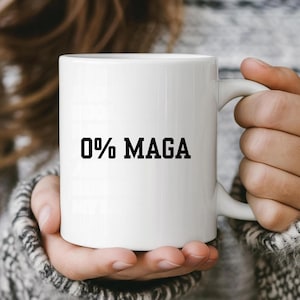 May include: White ceramic mug with the text "0% MAGA" in black block letters. The mug is held by a person wearing a gray sweater. The mug has a curved handle.