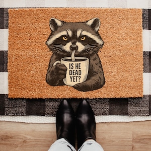 May include: A rectangular doormat with a raccoon illustration holding a coffee mug. The mug reads "IS HE DEAD YET?" The mat is brown with a textured surface, placed on a black and white checkered rug. Black boots are visible at the bottom.