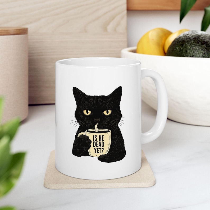 May include: White ceramic mug featuring a black cat illustration holding a coffee cup. The mug has the text "IS HE DEAD YET?" printed on the cup. The cat has yellow eyes and is set against a white background.