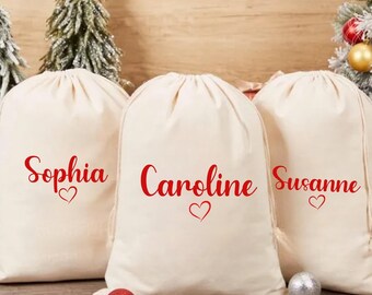 May include: Three off-white drawstring gift bags with red text that reads "Sophia", "Caroline", and "Susanne", each with a heart symbol. The bags are on a wooden surface, with Christmas decorations in the background.