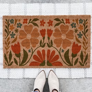 May include: A rectangular doormat with a floral design in shades of brown, orange, and green. The design features large flowers, tulips, and leaves. The mat is made of a natural fiber material and is placed on a patterned rug.