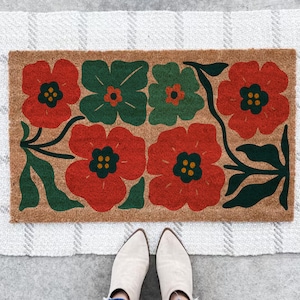 May include: A rectangular doormat with a natural coir fiber background. The mat features a floral design with red and green flowers. The flowers have yellow centers and are arranged in a symmetrical pattern. The mat is placed on a white and gray striped rug.
