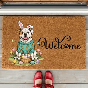 May include: A brown doormat with a smiling dog wearing bunny ears and a sweater with carrot designs. The dog is sitting in front of a basket of Easter eggs. The word "Welcome" is written in black script.