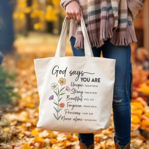 May include: Cream-colored canvas tote bag with the words "God says YOU ARE" and inspirational phrases like "Unique" and "Strong" in black script. Floral illustrations in yellow, pink, orange, and purple accent the text. The bag has sturdy handles.