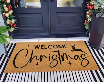 May include: A rectangular doormat with the words "WELCOME Christmas" in black script. A black silhouette of a reindeer is above the word "Christmas". The mat is tan and sits on a black and white striped rug.