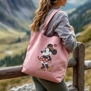 May include: A pink tote bag with a Minnie Mouse design. The cartoon character is wearing a red polka-dot dress and a matching bow. The bag has two shoulder straps and the Disney logo.