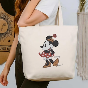 May include: Cream tote bag with a cartoon Minnie Mouse illustration in a red polka-dot dress and bow. The Disney logo is printed in black. The bag has two handles and is held by a person. The bag is made of a light-colored fabric.