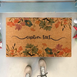 May include: A rectangular doormat with a floral design in shades of orange, green, and purple. The words "custom text" are written in black script across the center. The mat is made of a natural fiber material.