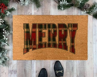 May include: A rectangular coir doormat with the word "MERRY" in large, block letters. The letters are filled with a red, green, and gold plaid pattern. The mat is on a light wood floor, with feet wearing black loafers visible.