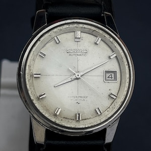 Peut inclure: Montre-bracelet automatique Seiko vintage avec un cadran argenté, des index rectangulaires et un guichet de date. La montre a un bracelet en cuir noir et les mots "SEIKO AUTOMATIC WATER PROOF" sont visibles sur le cadran.