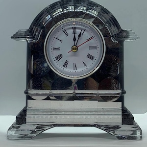 May include: A decorative clock with a clear, faceted arch and base. The clock face is white with black Roman numerals and red hands. The clock's body is a dark, speckled material.