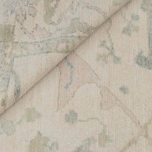 Op de afbeelding: Close-up van een opgerolde, crèmekleurige vloerkleed met een bloemenpatroon in gedempte tinten groen, roze en blauw. Het ontwerp van het vloerkleed bevat gestileerde bloemen- en bladmotieven, waardoor een zacht, getextureerd uiterlijk ontstaat.