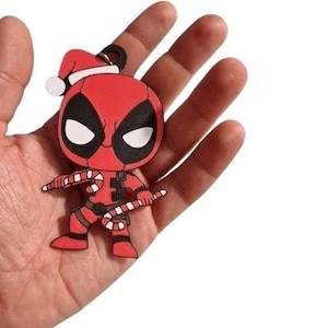 Può includere: Un portachiavi rosso di Deadpool con un cappello da Babbo Natale e bastoncini di zucchero. Il portachiavi presenta gli occhi bianchi e neri caratteristici del personaggio ed è tenuto in mano su sfondo bianco. Un articolo festivo natalizio.
