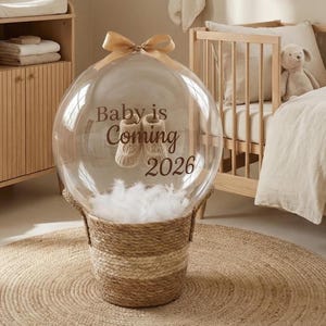 Op de afbeelding: Een doorzichtige bolvormige ballon staat in een geweven beige mand gevuld met witte veren. De ballon heeft een beige strik aan de bovenkant en bruine tekst met Baby is Coming 2026, met een paar kleine beige gebreide babyslofjes erin.