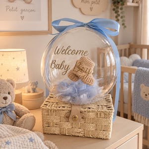 Peut inclure: Un ballon sphérique transparent avec le texte Welcome Baby Boy imprimé en noir, contenant des plumes bleu clair et une paire de petits chaussons tricotés beiges. Le ballon est noué avec un ruban bleu clair et posé sur un panier rectangulaire tressé.