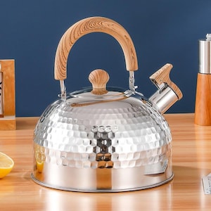 May include: A stainless steel tea kettle with a hammered texture. It features a wooden handle, lid knob, and spout accent. The kettle sits on a wooden surface with a blue background.