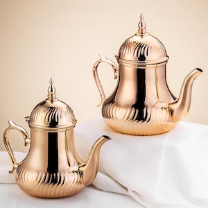 May include: Two polished gold-colored teapots with ornate lids and curved spouts. The teapots have a textured, ribbed design on the lower portion and a smooth, reflective surface. The teapots are placed on a white surface.