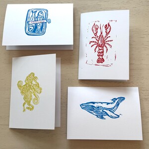 May include: Four white folded greeting cards featuring block print illustrations of sea creatures. One card shows a blue tin of sardines with the text "sardines", one a red lobster, one a yellow octopus, and one a blue whale in a side profile.