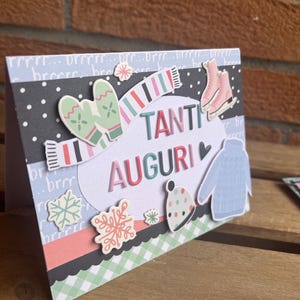 Può includere: Biglietto di auguri fatto a mano a tema invernale. Il biglietto presenta un ovale bianco con le parole "TANTI AUGURI" a lettere colorate. Elementi decorativi: guanti, sciarpa, pattini, cappello, maglione e fiocchi di neve.