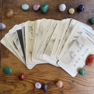 May include: A deck of oracle cards with a landscape design and text, fanned out on a wooden surface. Various colorful polished stones, including pink, green, and purple, are scattered around the cards.