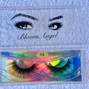 Könnte beinhalten: Eine transparente Kunststoffhülle mit einem Paar schwarzer falscher Wimpern. Der Deckel der Hülle zeigt eine Zeichnung von Augen und den Text "Bloom Angel". Der Boden der Hülle hat einen holografischen Hintergrund mit dem Etikett "H8".