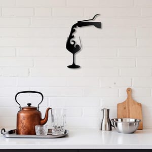 Może przedstawiać: Czarna metalowa grafika ścienna przedstawiająca butelkę wina wlewaną do kieliszka. Dzieło sztuki jest zamontowane na białej ceglanej ścianie. Na blacie kuchennym poniżej znajduje się miedziany czajnik, szkło i drewniana deska do krojenia.