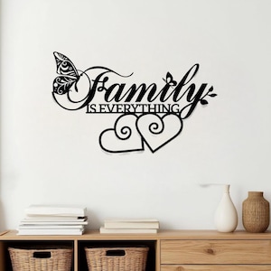 May include: Black metal wall art with the words "Family is Everything" and two heart shapes. A butterfly and leaves are incorporated into the design. The art is displayed on a white wall above a wooden cabinet.