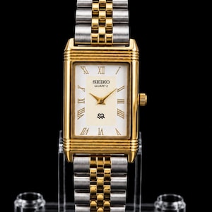 Peut inclure: Une montre-bracelet rectangulaire Seiko Quartz avec un boîtier doré et un bracelet à maillons en métal bicolore or et argent. Le cadran blanc présente des chiffres romains dorés, des aiguilles dorées et un petit logo noir à six heures.