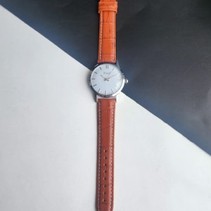 May include: A silver wristwatch with a white face and the brand name "Piaget" printed on it. The watch has a brown leather band with a textured pattern. The watch is displayed against a black and white background.