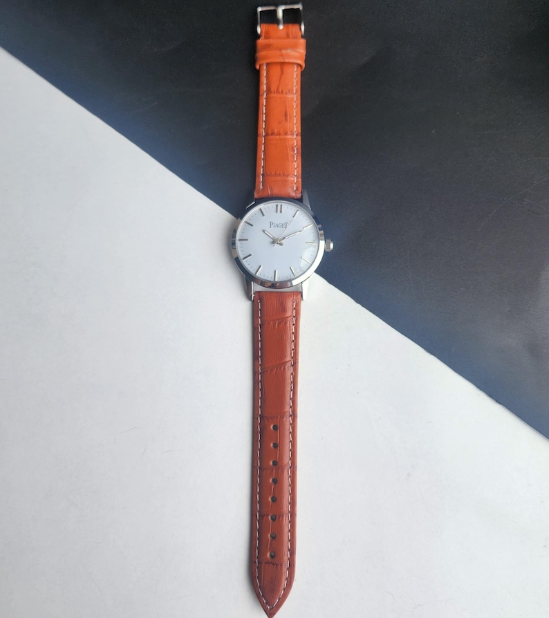 May include: A silver wristwatch with a white face and the brand name "Piaget" printed on it. The watch has a brown leather band with a textured pattern. The watch is displayed against a black and white background.