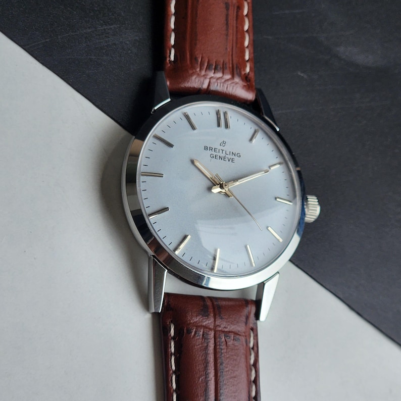 Op de afbeelding: Een zilveren Breitling Gen&egrave;ve polshorloge met een bruine leren band. Het horloge heeft een lichtgekleurde wijzerplaat met zilveren uurmarkeringen en gouden wijzers. De band is donkerbruin met witte stiksels.