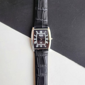 Puede incluir: Reloj Seiko Quartz rectangular con esfera negra y caja plateada. El reloj presenta números romanos y una correa de cuero negro con un patrón de cocodrilo.