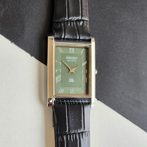 Pode incluir: Um rel&oacute;gio Seiko Quartz retangular com mostrador verde e ponteiros dourados. O rel&oacute;gio tem uma caixa prateada e uma pulseira de couro preta. Os algarismos romanos marcam as horas. O mostrador tem o logotipo Seiko Quartz.