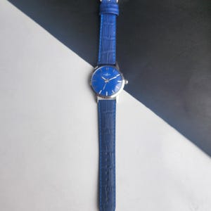 Puede incluir: Un reloj de pulsera azul con caja plateada y correa de cuero azul a juego. La esfera del reloj es de un azul vibrante con manecillas e indicadores plateados. La correa tiene una textura de cocodrilo.