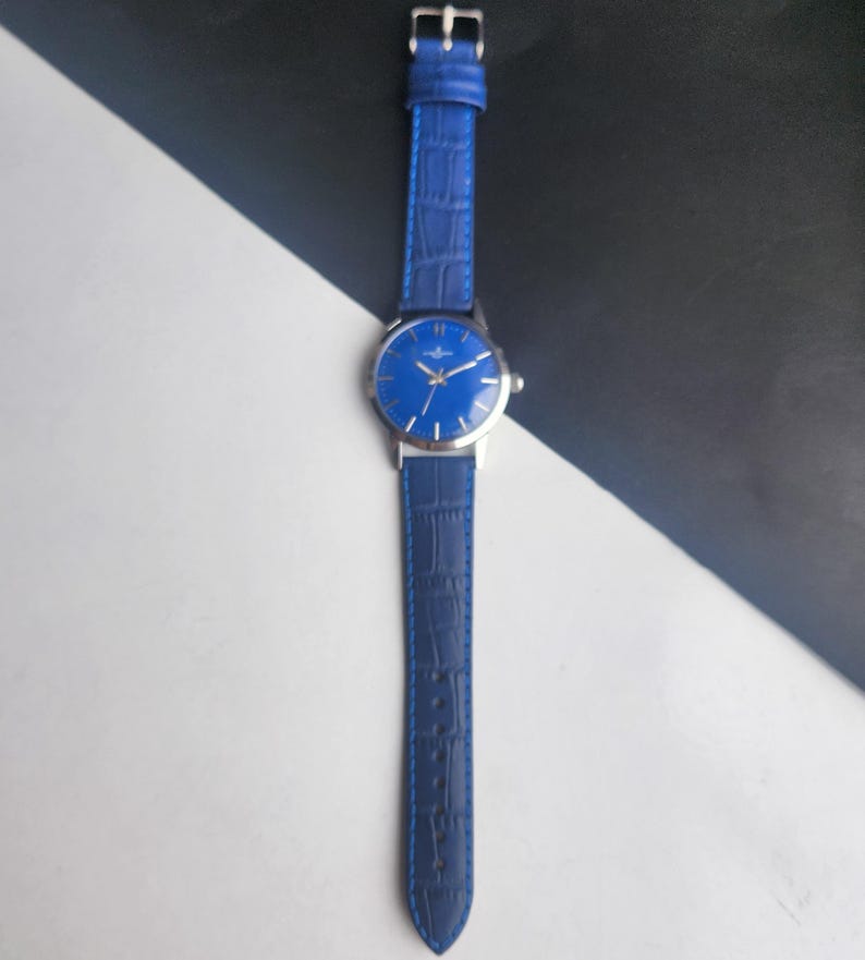 Puede incluir: Un reloj de pulsera azul con caja plateada y correa de cuero azul a juego. La esfera del reloj es de un azul vibrante con manecillas e indicadores plateados. La correa tiene una textura de cocodrilo.
