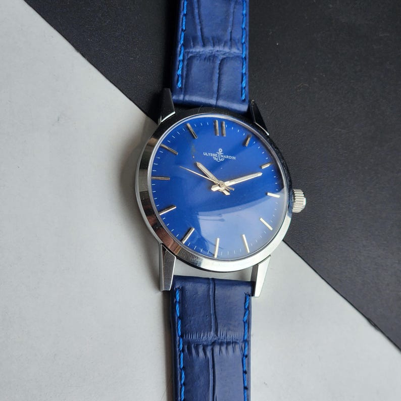Puede incluir: Un reloj de pulsera con esfera azul brillante y caja plateada. El reloj tiene una correa de cuero azul con un patr&oacute;n de cocodrilo. La esfera del reloj tiene agujas e indicadores de hora plateados. La marca "ULYSSE NARDIN" se muestra en la esfera.