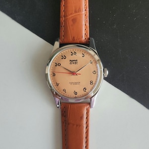 Könnte beinhalten: Eine Vintage-Armbanduhr mit hellbraunem Zifferblatt und braunem Lederarmband. Das Zifferblatt zeigt arabische Ziffern und Text in einer Fremdsprache. Die Uhr hat ein silberfarbenes Gehäuse.