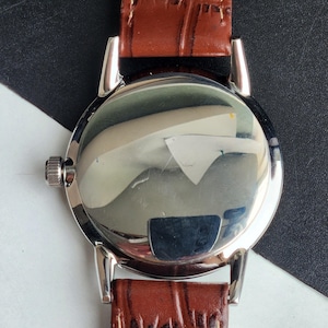 Op de afbeelding: Close-up van de achterkant van een zilveren polshorloge met een bruine leren band. De wijzerplaat van het horloge is reflecterend en toont een vervormd beeld. De band is rijk bruin van kleur en het horloge heeft een zilveren kroon.