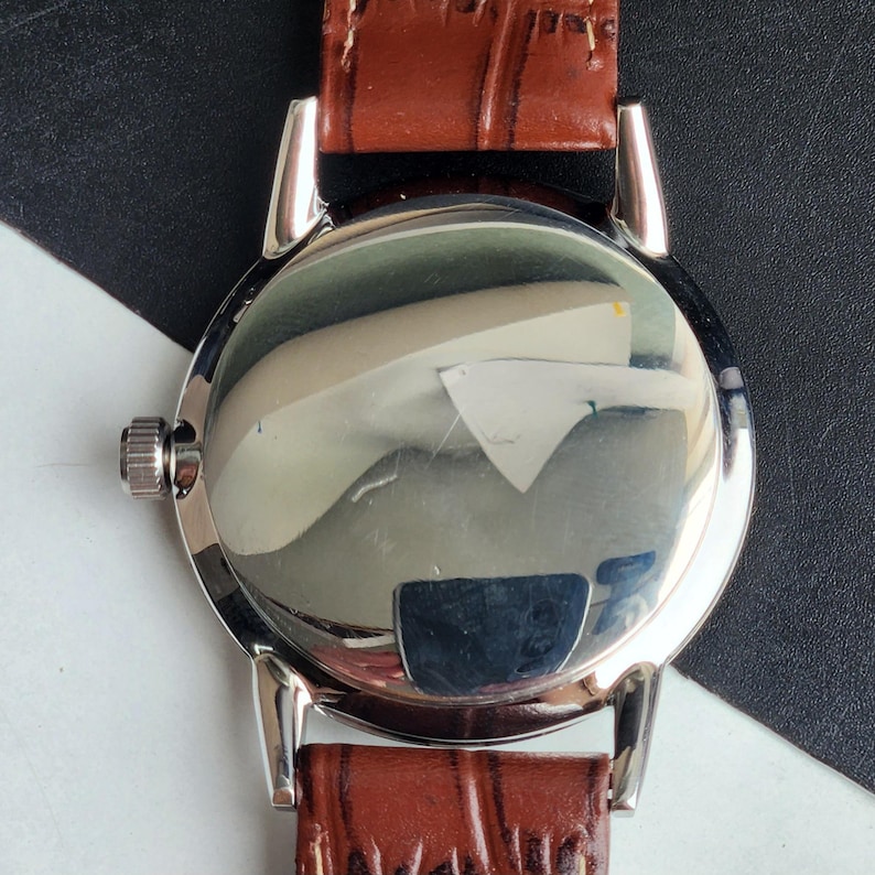 Op de afbeelding: Close-up van de achterkant van een zilveren polshorloge met een bruine leren band. De wijzerplaat van het horloge is reflecterend en toont een vervormd beeld. De band is rijk bruin van kleur en het horloge heeft een zilveren kroon.