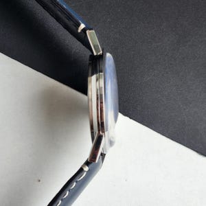 May include: A silver wristwatch with a blue face and a black leather strap. The watch has a round face and a modern design. The strap is stitched with white thread.