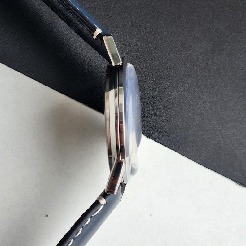 May include: A silver wristwatch with a blue face and a black leather strap. The watch has a round face and a modern design. The strap is stitched with white thread.