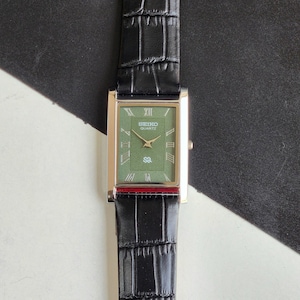 Pode incluir: Um rel&oacute;gio Seiko Quartz retangular com mostrador verde e caixa dourada. O rel&oacute;gio tem uma pulseira de couro preta com padr&atilde;o de crocodilo. O mostrador exibe algarismos romanos e o logotipo Seiko Quartz.