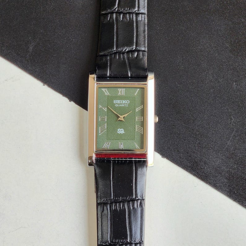 Pode incluir: Um rel&oacute;gio Seiko Quartz retangular com mostrador verde e caixa dourada. O rel&oacute;gio tem uma pulseira de couro preta com padr&atilde;o de crocodilo. O mostrador exibe algarismos romanos e o logotipo Seiko Quartz.