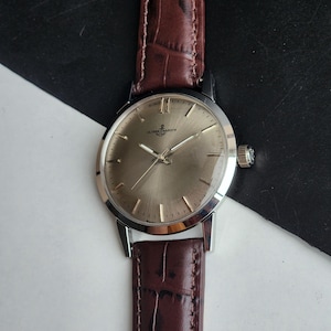May include: A silver-toned wristwatch with a brown leather band. The watch face is a light gold color with silver hands and markers. The brand name "ULYSSE NARDIN" is visible on the watch face. The band is a dark brown with a textured pattern.