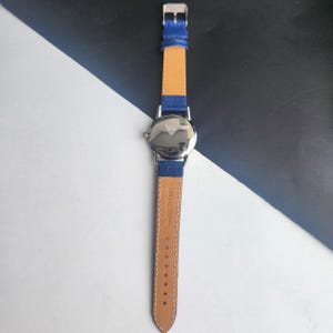 Puede incluir: Un reloj de pulsera plateado con una correa de cuero azul y un respaldo de cuero marr&oacute;n claro. El reloj tiene una esfera redonda y un cierre de hebilla. La correa est&aacute; marcada "GENUINE LEATHER".