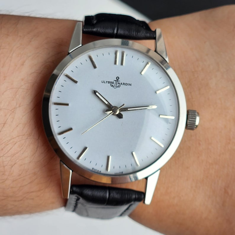 May include: A silver-toned wristwatch with a light-colored face and black leather band. The watch face features the brand name "ULYSSE NARDIN" and a small anchor emblem. The watch has a round face with silver hour markers and hands.